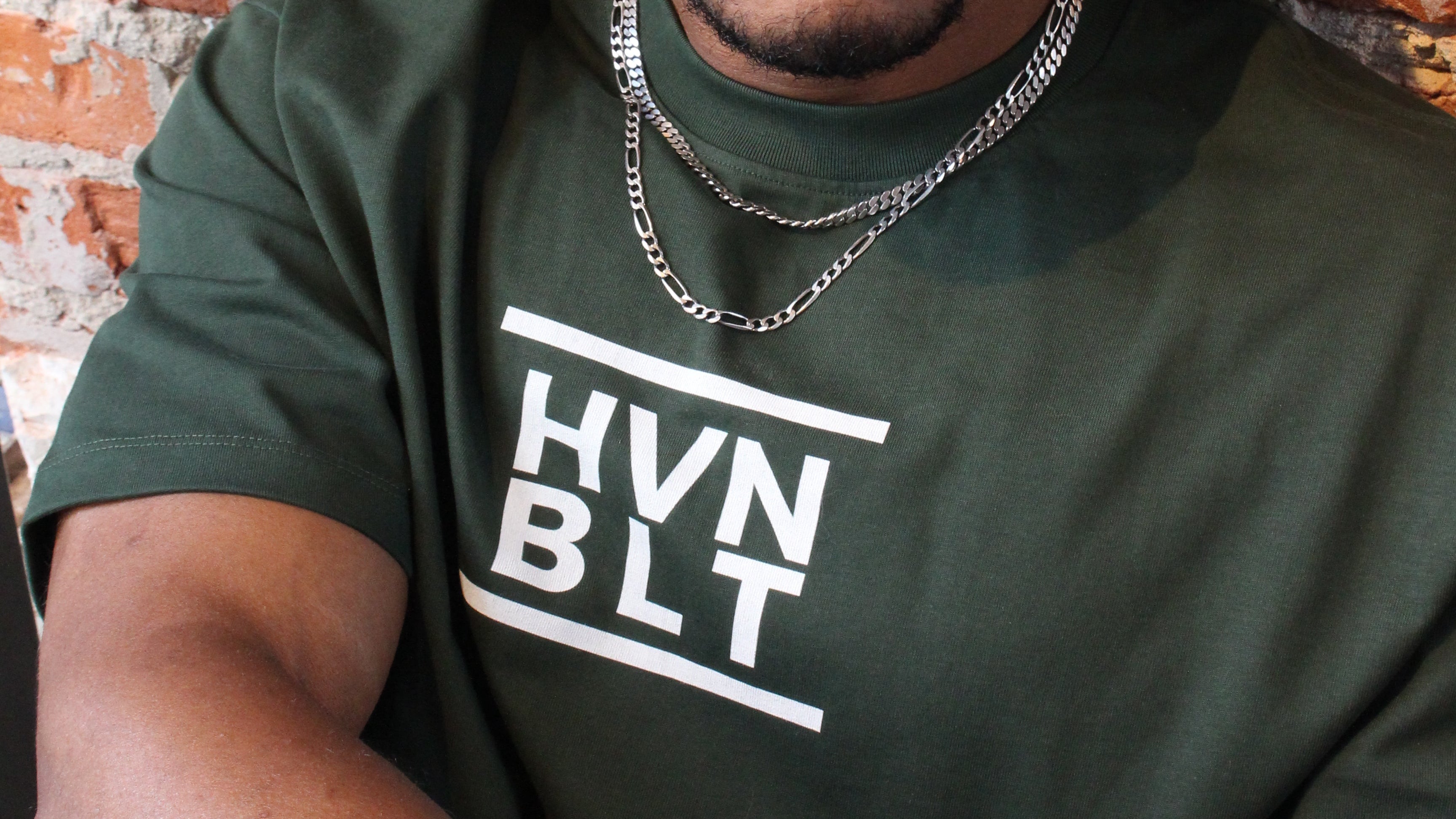 Man wearing a green t-shirt with 'HVN BLT' text against a brick wall.