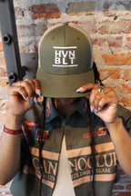 Person holding a green cap with 'HVNBLT' logo against a brick wall.